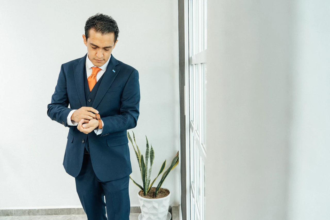 El novio con traje azul ajusta su reloj junto a la ventana